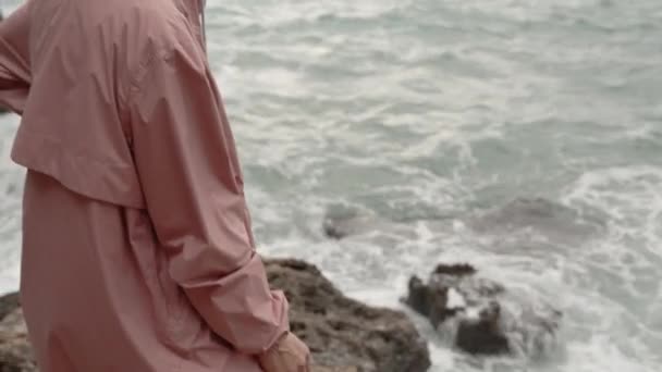 카메라는 날카로운 바위와 가장자리에 서있는 여자를 포착합니다 바다에 파도가있는 폭풍이있다 — 비디오
