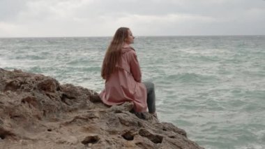 Uzun Saçlı Genç Kadın Denizde Bir Uçurumun Kıyısında Oturuyor ve Uzağa Bakıyor. Yalnızlık ve Endişe kavramı Denizdeki Fırtına gibi.