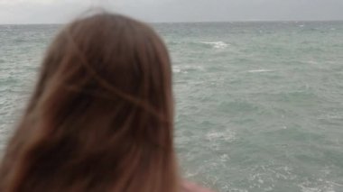 Genç Bir Kadının Başının Arkasından, Uzun Saçlı, Denizde Bir Uçurumun kenarında Duran ve Yaklaşan Fırtınadaki Mesafeye Bakan. Kamera Dalgalara Odaklanıyor.