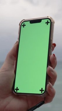Dikey video. Dişi El, Deniz Arkaplanı ve Bulutlu Hava Durumu 'na Karşı Boş Yeşil Ekran ve İşaretleyiciler ile Bir Cep Telefonu Tutar.