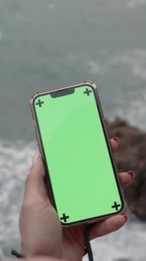 Dikey video. Fırtınalı Deniz kıyısında Büyük Dalgalar ve Bulutlu Hava Durumu ile Bir Uçurumun Kıyısında Dururken Elinde Yeşil Ekranlı Akıllı Bir Telefonla Kadın Başının Arkasından Görüntü.