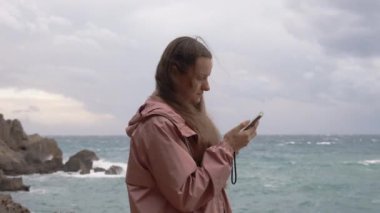 Deniz kenarındaki fırtınada genç bir kadın kayalıklarda duruyor, telefonunda bir şeye bakıyor. Hava bulutlu, pembe bir yağmurluk giyiyor..