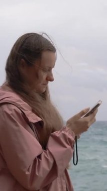 Dikey video. Deniz kenarındaki fırtınada genç bir kadın kayalıklarda duruyor, telefonunda bir şeye bakıyor. Hava bulutlu, pembe bir yağmurluk giyiyor..