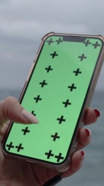 Dikey video. Yeşil Ekran ve İzleme İşaretli Akıllı Telefon, Ekranda Büyük Parmaklı Çapraz Kadın. Deniz Dalgaları ve Kayaların Arkaplanı 'na karşı, Akşam Işığı.