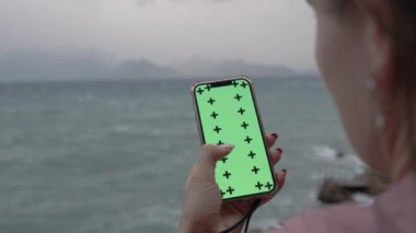 Deniz kenarındaki Bulutlu Akşamın Arkaplanına Karşı Yeşil Ekranlı Akıllı Telefondan Bir Şey Arayan Bir Kadının Başının Arkasından Görünümü. Arka planda Dağlı Yüksek Kayalıklar.