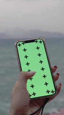 Dikey video. Deniz kenarındaki Bulutlu Akşamın Arkaplanına Karşı Yeşil Ekranlı Akıllı Telefondan Bir Şey Arayan Bir Kadının Başının Arkasından Görünümü. Arka planda Dağlı Yüksek Kayalıklar.