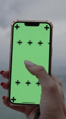 Dikey video. Bir kadının elinde yeşil ekran ve takip işaretleri olan akıllı bir telefon, denizdeki bulutlu ve yağmurlu havanın arka planına karşı yan yana kayıyor..