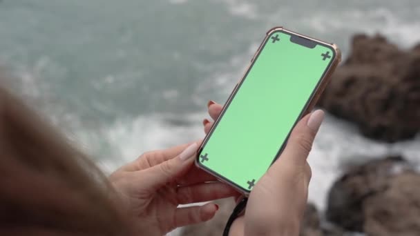 妇女手持带有绿色屏风的电话 跟踪标识 站在高高的悬崖上 身后的波浪冲击着岩石 — 图库视频影像