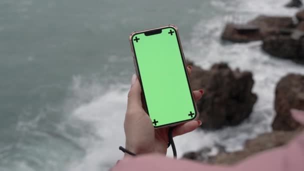 一只女手拿着带绿色屏风的电话 高高地站在悬崖边上 在下面 波浪冲击岩石 喷出水花 — 图库视频影像