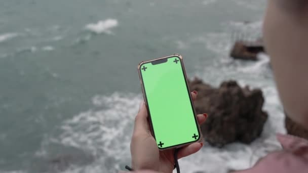 当她站在狂风暴雨和阴云密布的大海边的时候 从女性头后面看她拿着一个带着绿色屏风的智能手机 — 图库视频影像