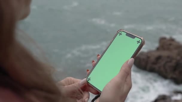 带着绿色屏风和跟踪标志的智能手机被站在海边悬崖边的长发女人握住 多云天气及大浪 — 图库视频影像