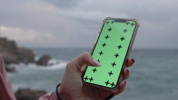 带有绿色屏幕和跟踪标记的智能手机 女人带着一个大手指在屏幕上来回穿梭 在海浪和岩石的背景下 — 图库视频影像