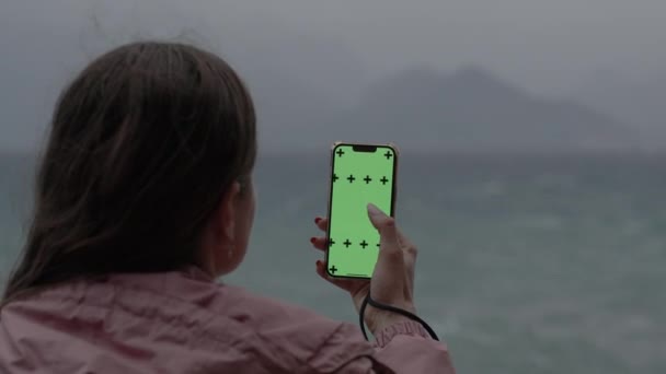 从后脑勺往外看 一个女人在绿色屏幕上用一个大手指在饲料里打转 把她的视频插入到标志上 在背景中 有大海 山脉和乌云天气 — 图库视频影像