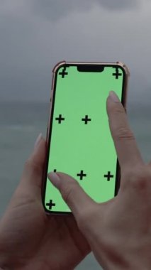 Dikey video. Yeşil ekranlı akıllı telefon ve denizin arka planına işaret koyan işaretler. Bir kadın ekranda yakınlaştırma hareketi yapar, bir gezginin haritasını görme ya da telefondaki bir resme yakınlaşma kavramı..