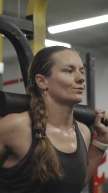 Dikey video. Smith makinesinde çömelme yapan genç bir kadın. Örgülü uzun saçları terli omuzlarında duruyor..