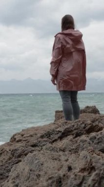 Dikey video. Yalnız genç bir kadın denizin bulutlu havasında bir uçurumda duruyor ve dağlara bakıyor. Rüzgarlı.