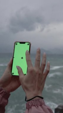 Dikey video. Dişi Eller Akıllı Telefonu Yeşil Ekran ile Tutar ve İşaretler Bir Mockup İçin, Videolarını Ekler. Seyahat ya da Fotoğraf Haritasında Ölçekleme kavramı. Bulutların Arkaplanı ve Denizde Fırtınaya Karşı.