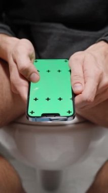 Dikey video. Pantolonunu indirmiş Bir Adam Tuvalette Oturuyor ve Yeşil Ekran ve İzleme İzleri ile akıllı bir telefondaki fotoğraflara bakıyor. Boşluğu Kopyalamak için Yeşil Ekranı kullan.