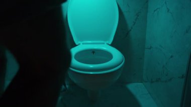 Adamın biri loş mavi ışıkla karanlık bir banyoya giriyor, şortunu çıkarıyor ve tuvalete oturuyor..