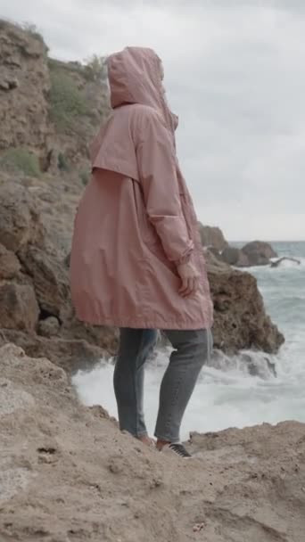 バーティカルビデオ ピンクのレインコートのシャープロックの女性のスタンドは 彼女の横の石に大きな波が衝突するのを見ています 海での嵐 海の嵐 — ストック動画