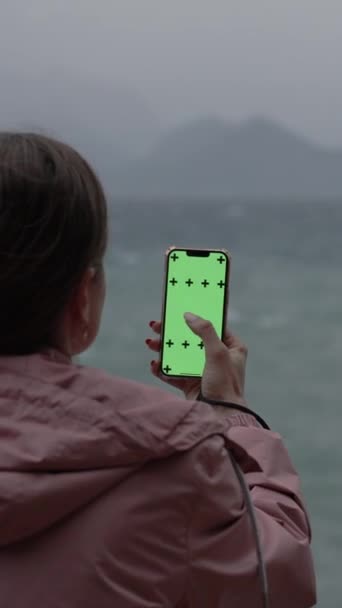 垂直录像 在阴云密布的天气里 一位穿着粉色外套的妇女在带绿色屏风的智能手机上翻阅照片 并在山脉背景下跟踪标识 — 图库视频影像