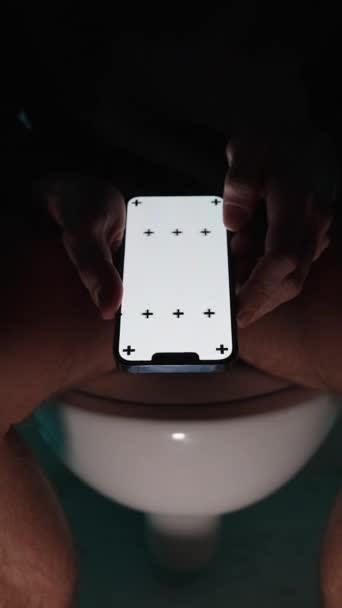 垂直录像 在黑暗中 一个男人在厕所里用一个空的白色屏幕在智能手机里打滚 他把它的特写 并继续翻阅页面 显示给你看 使用白色屏幕作为复制空间 — 图库视频影像