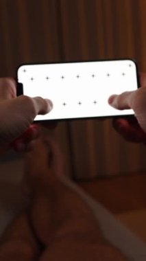 Dikey video. Bakış açısı. Smartphone 'u tutan ve yatakta yatan bir adam. Beyaz Ekran ve İzleme İşaretleri 'nin bir düzenine sahip akıllı telefon.