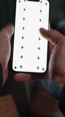 Dikey video. Bakış açısı. Adam Yatağın Üzerinde Karanlıkta Yatıyor, Beyaz Ekran Düzeni ile Akıllı Telefondan Kaydırıyor. 