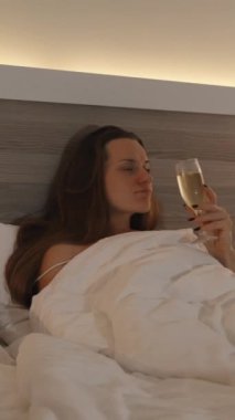 Dikey video. Gecenin geç saatlerinde, Yatakta Genç Bir Kadın Şampanyasını yudumluyor ve TV seyrediyor. Kamera yavaşça geriye doğru hareket ediyor. Parlak Yatak Odası İçi.