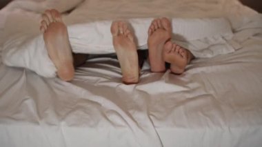 Battaniyenin altında birbirini okşayan bir kadın ve bir erkeğin bacakları. Yatak odasında TV ışığı ve akşam dekorasyonu.