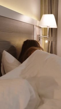 Dikey video. Yatak odasındaki akşam atmosferinde, yalnız bir kadın komodinden bir bardak şampanya alır ve televizyon izlerken içer..