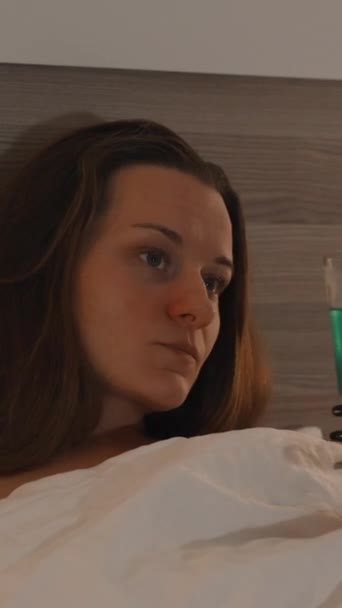 バーティカルビデオ カクテルをシッピングする若い女性のクローズアップ ベッドのカバーの下に横たわり ベッドの前にテレビを見る — ストック動画