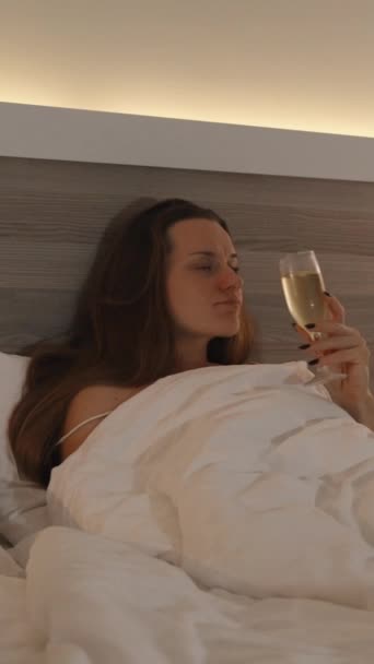 バーティカルビデオ 夜遅く ベッドに座った若い女性がシャンパンとテレビを見る カメラはゆっくりと後方に移動する 明るいベッドルームインテリア — ストック動画