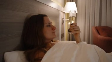 Yatakta genç bir kadının portresi, kamera yakınlaşır, şampanyasını yudumlarken yakın plan bir fotoğraf çeker. TV ışığı arka planda titreşiyor..