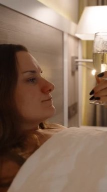 Dikey video. Yatakta genç bir kadının portresi, kamera yakınlaşır, şampanyasını yudumlarken yakın plan bir fotoğraf çeker. TV ışığı arka planda titreşiyor..