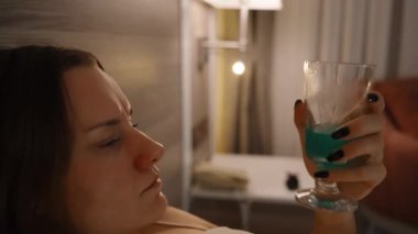Yatakta bir kadının mavi bir kokteyli yudumlarken, yatak odasındaki lambalardan loş bir akşam ışığında televizyon izlerken..