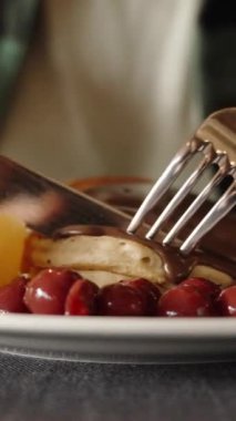 Dikey video. Çikolata, kiraz meyvesi ve şeftali kaplı bir tabağın yakın çekimi. Bir kadın bir parça keser ve yer. Restoranda kahvaltı..