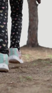 Dikey video. Sunny Forest 'ta ailesiyle yürüyen küçük bir kızın bacakları. Yavaş Çekimde Aşağıdan Görüntüle.