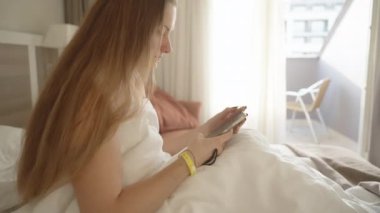 Beyaz bir otel yatağında bilekliğinde sarı bir bileklik takan genç bir kadın. Telefonunda bir şeye bakıyor, muhtemelen oda servisine sipariş veriyor. Güneş ışınları balkon kapısında parlıyor..