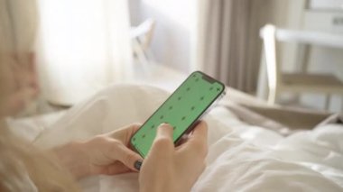 Yeşil Ekran Modeli ve İzleme İşaretleri olan akıllı telefon, beyaz bir yatakta otururken sosyal medyada gezinen bir kadın..