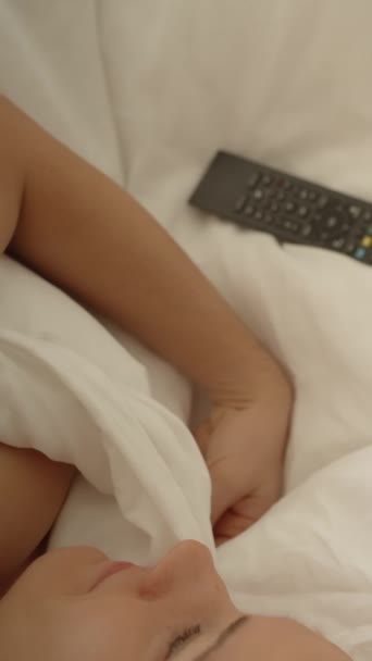 垂直录像 早上躺在白床里的年轻女人用遥控装置打开电视的头像 — 图库视频影像