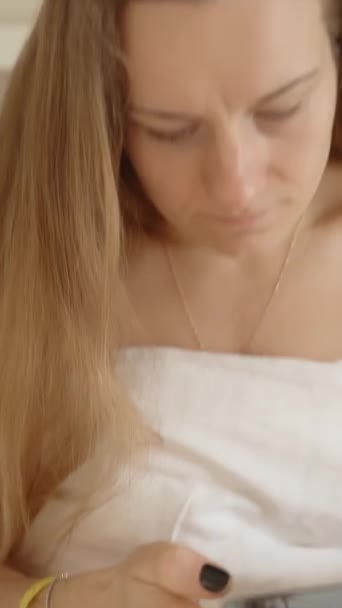 垂直录像 一个年轻的女人坐在一张白色的床上 看着她手机上的某个东西 酒店房间概念 白色内政 — 图库视频影像