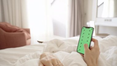 Kadın Eller Akıllı Telefonu Yeşil Ekran ile Tutar ve Parmakla Ekrandan Geçer. Güneşli bir sabahta, beyaz bir yatakta uzanıyor. Boş bir ekranın maketi ile..