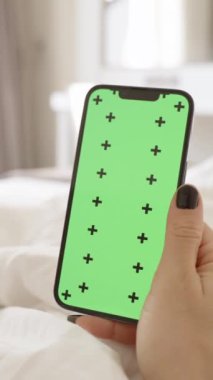 Dikey video. Beyaz Yataktaki Kadın Eli 'ndeki Smartphone' da Chroma Key Mockup. Parlak Yatak Odası İçi.