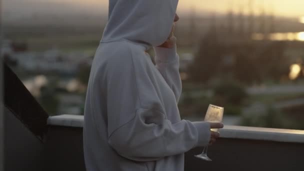 日落时 一个穿着白色帽衫的女人站在阳台上喝香槟 在远方 群山和船只 — 图库视频影像