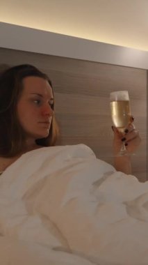 Dikey video. Genç bir kadın, akşam geç saatte yatakta yatarken şampanya içiyor, bir lambayla aydınlatılıyor ve televizyon seyrediyor..