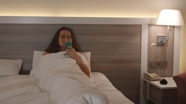 Genç Bir Kadın Beyaz Yatakta Uzanıp Akşam Ortamında Televizyon izlerken Kokteylden Hoşlanıyor.