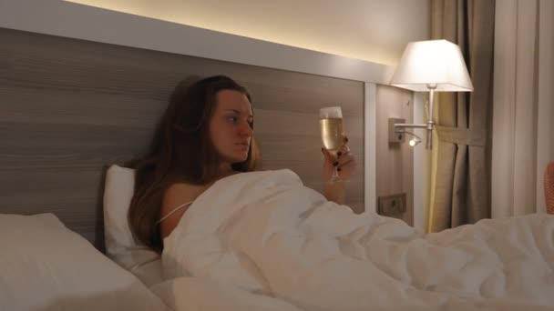 一个年轻的女人晚上躺在床上喝香槟 被灯照亮 看电视 — 图库视频影像