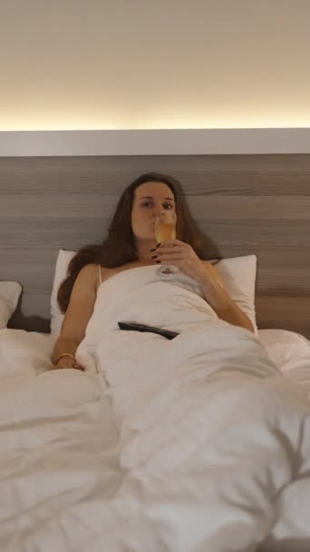 垂直录像 夜晚的气氛 灯照亮了床 女人躺在床上 喝香槟 看电视 — 图库视频影像