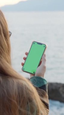 Uzun Saçlı Bir Kadının Arkasından, Elinde Akıllı Telefonun Yeşil Ekranına Bakış. Gün batımında Deniz ve Dağların Arkaplanına karşı.